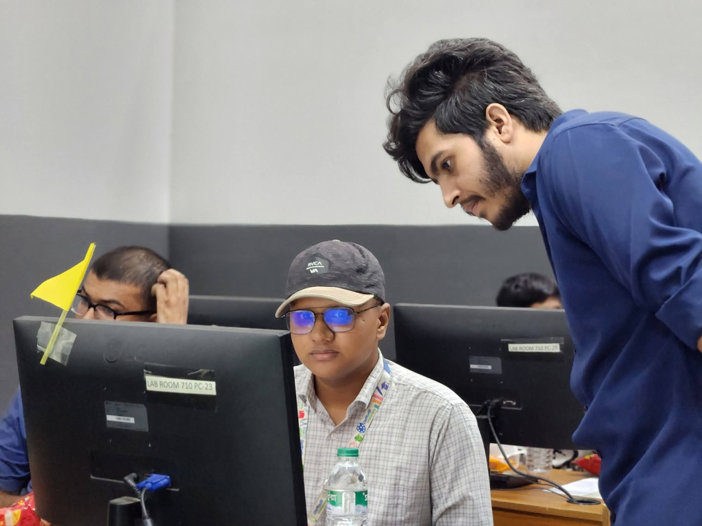 Shafim leaning over to assist and guide a teammate working at a computer lab desk during a hackathon